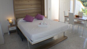 Una cama bien hecha con cabecera de madera, una mesita de noche con lámpara, y una silla junto a una mesa de comedor con vistas al exterior.