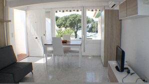 Una sala de estar moderna con televisor, un comedor con sillas y una mesa, y vista a árboles a través de la puerta abierta.