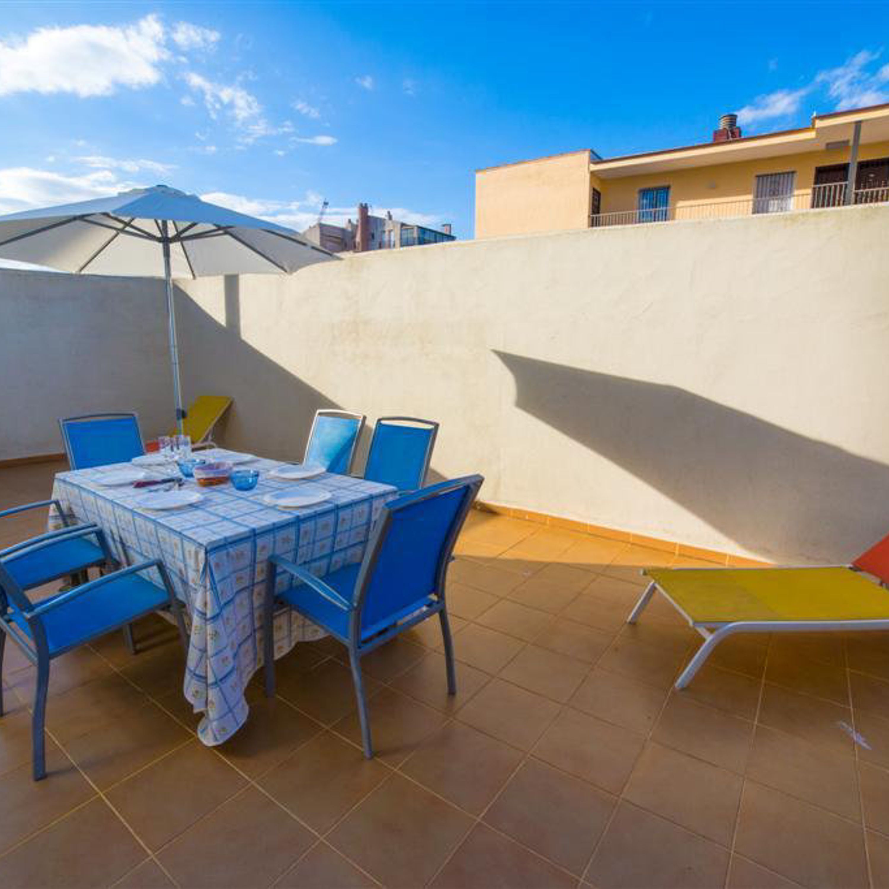 A patio with a table set for four, blue chairs, a yellow lounge chair, and a large umbrella.