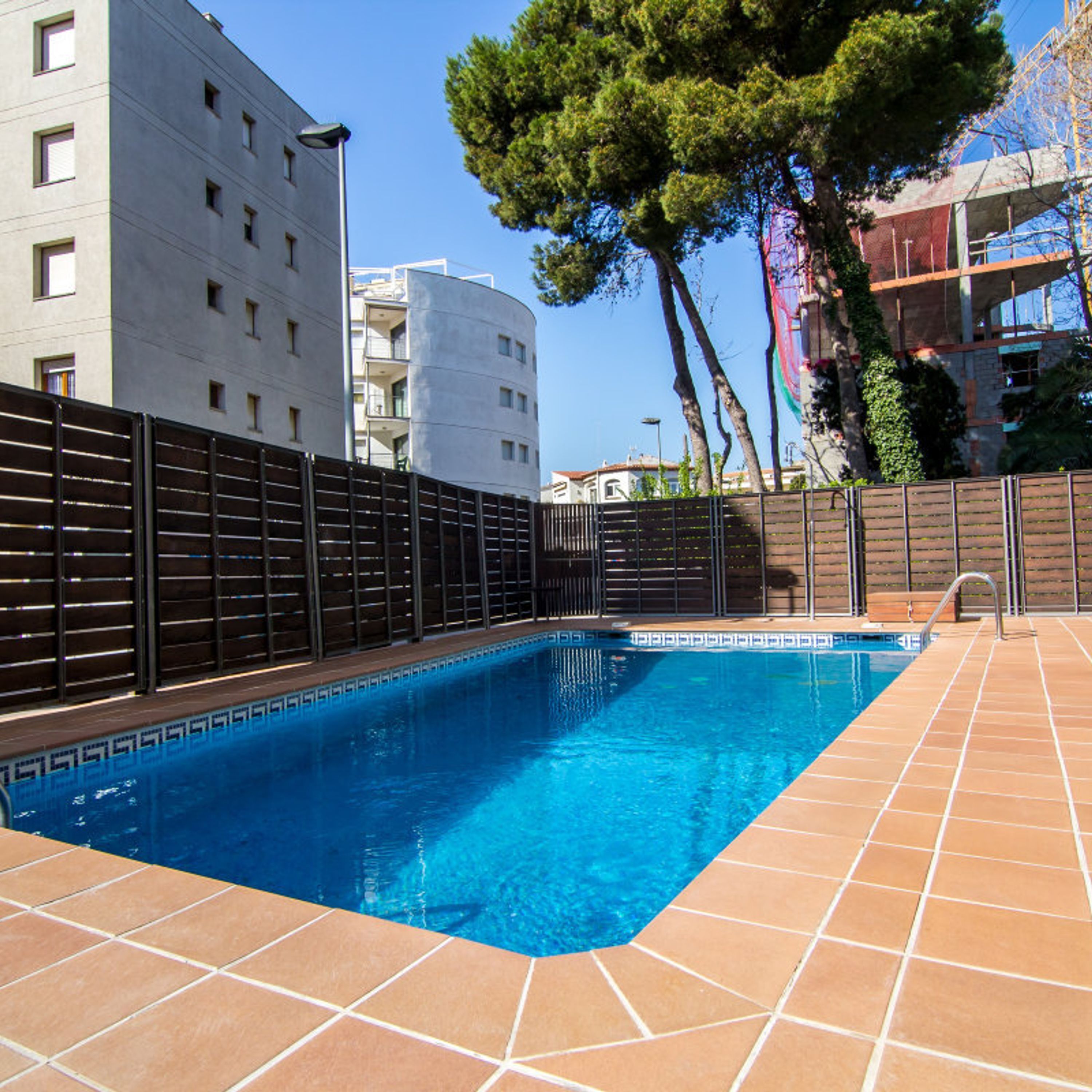 Une piscine rectangulaire avec une terrasse carrelée, entourée d’une petite clôture et d’un arbre.