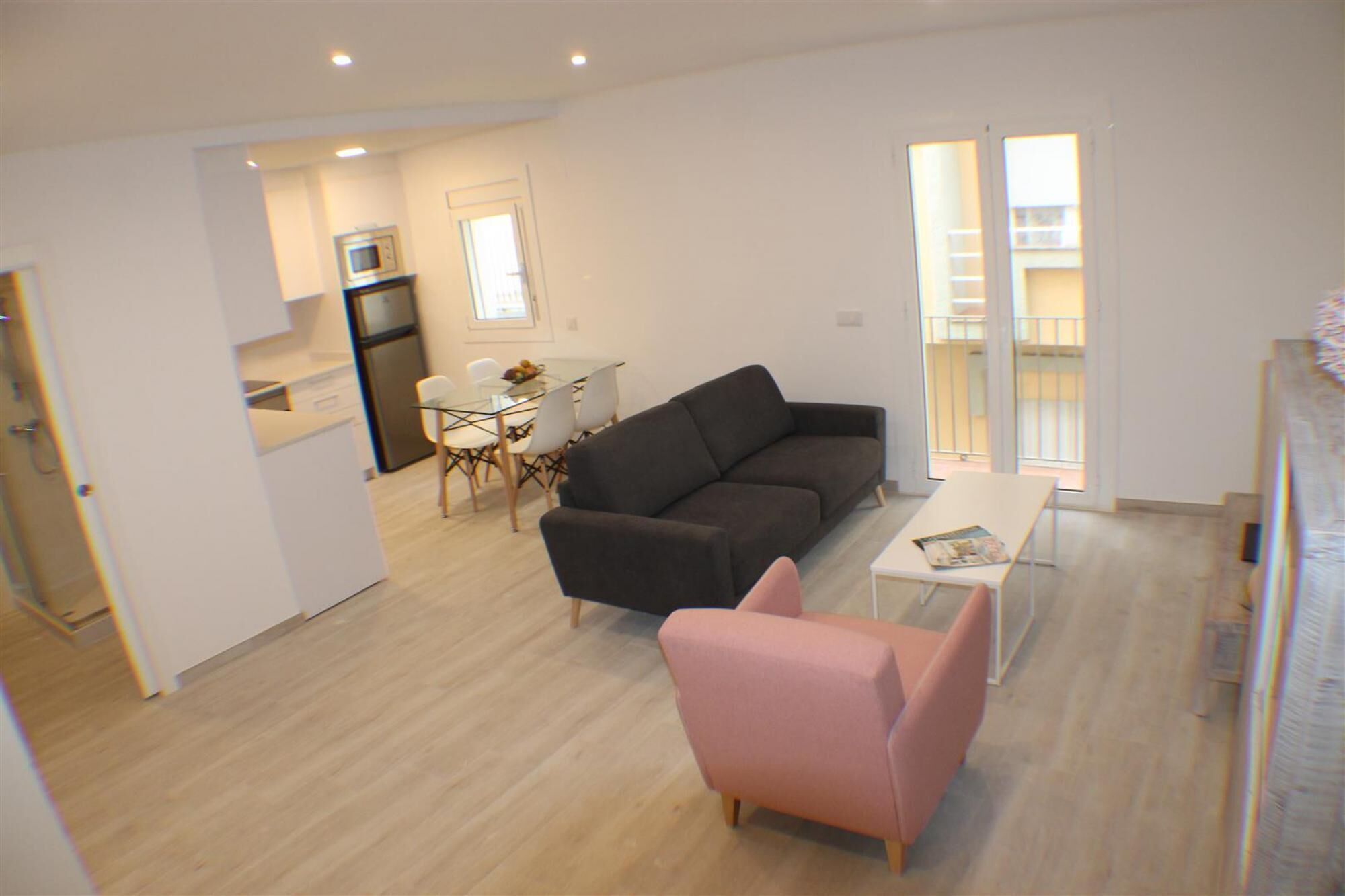 A modern living room with a dark sofa, a pink armchair, and a glass dining table. There is a kitchen area with a refrigerator and a microwave. The room has a wooden floor and large windows with a view of a balcony.