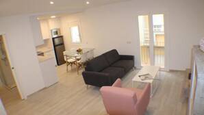 A modern living room with a dark sofa, a pink armchair, and a glass dining table. There is a kitchen area with a refrigerator and a microwave. The room has a wooden floor and large windows with a view of a balcony. - Cosconilles 1 in Roses (Roses)