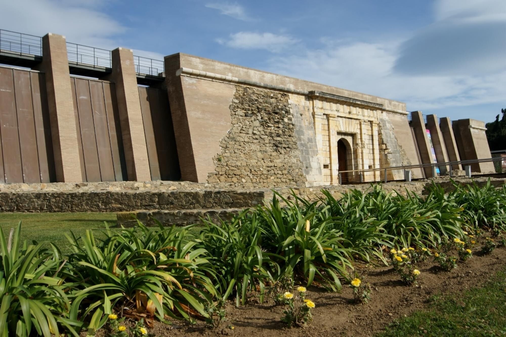 A building with a stone facade, a metal gate, and a paved area with plants and flowers.