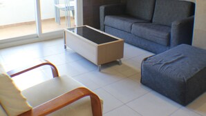 Un espacio habitable pequeño con un sofá gris, un pequeño reposapiés, una mesa de centro de vidrio y una silla blanca. Una puerta corrediza de cristal da a un área exterior con una silla azul.