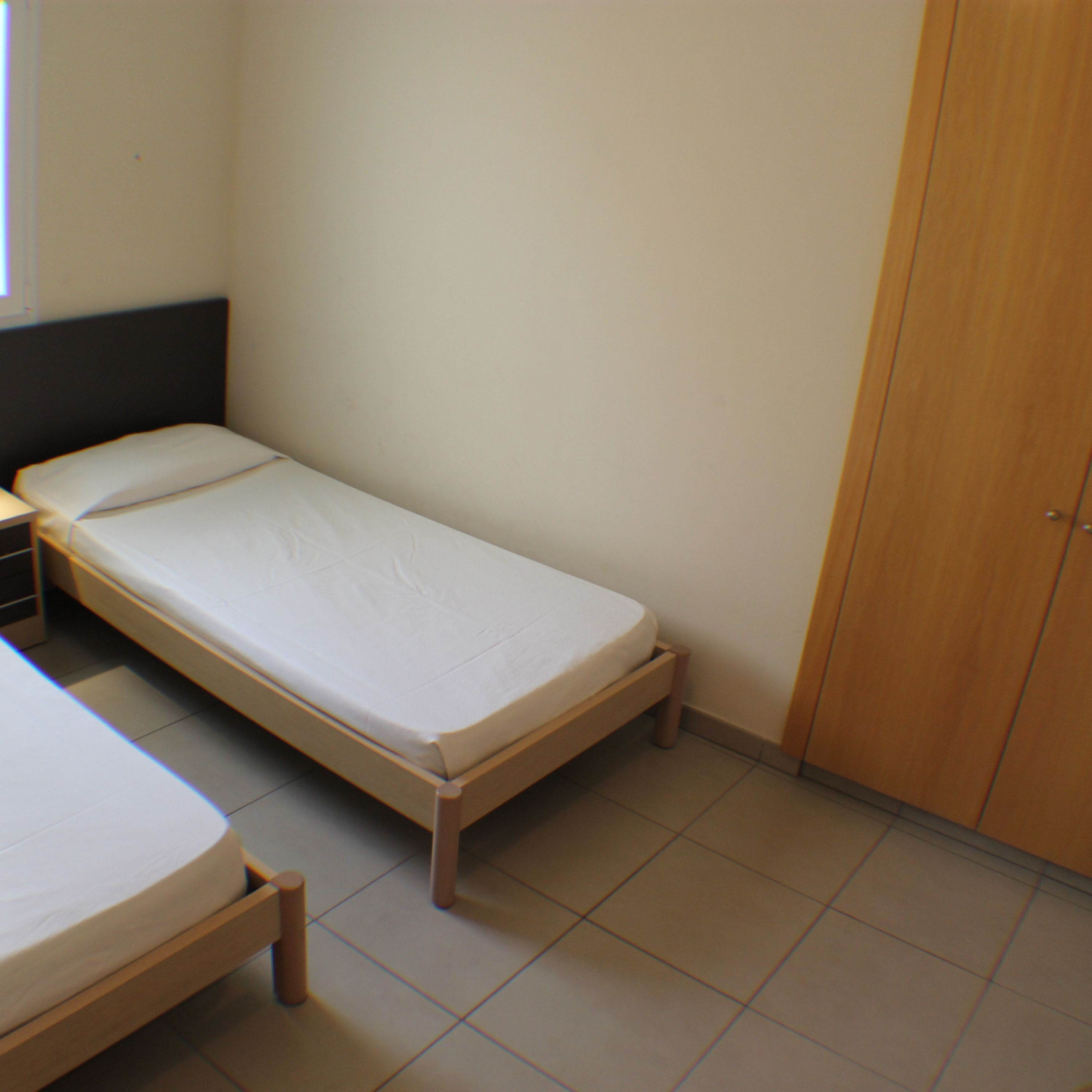A single bed with a wooden frame and a white mattress in a small room with tiled flooring.