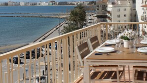 Un balcon avec vue sur la mer, une table à manger dressée avec des assiettes et des verres, et une rangée de voitures garées.