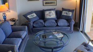 Un salon avec un canapé bleu, une table basse en verre et des œuvres d’art sur le thème de la marine, encadrées et accrochées au mur.