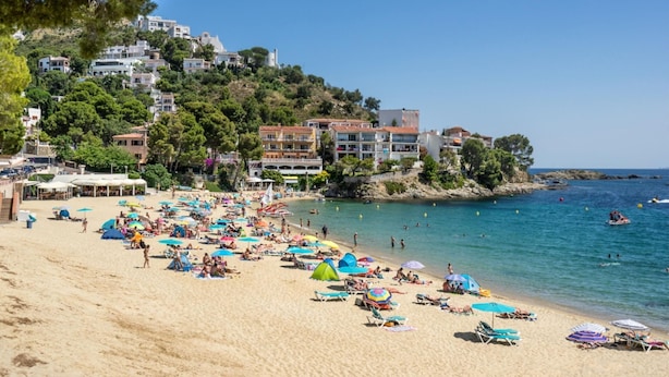 Egy strand, ahol rengeteg napernyő és napozóágy található, a tenger kristálytiszta és kék, a háttérben pedig egy domboldalra épült épületek láthatók.