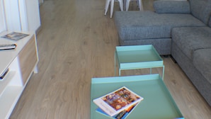 A modern living room with a grey sofa, a dining table with chairs, and a kitchen in the background.