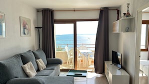 A modern living room with a grey sofa, a small TV, and a balcony with a view of the sea.