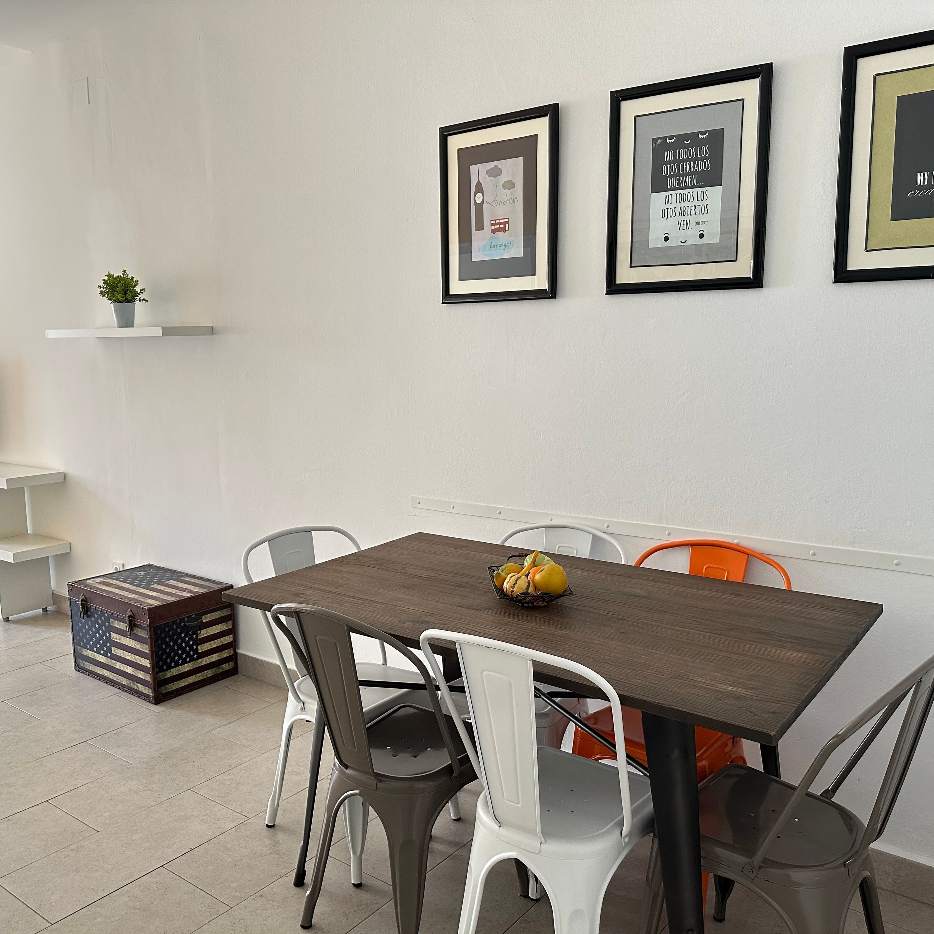 Un comedor con una mesa de madera, sillas blancas y grises, y una pequeña planta en un estante blanco.
