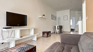 A living room with a grey sofa, a flat-screen TV on a white stand, a small table with an American flag design, and a dining area in the background. - Joan Badosa 1 B in Roses (Roses)