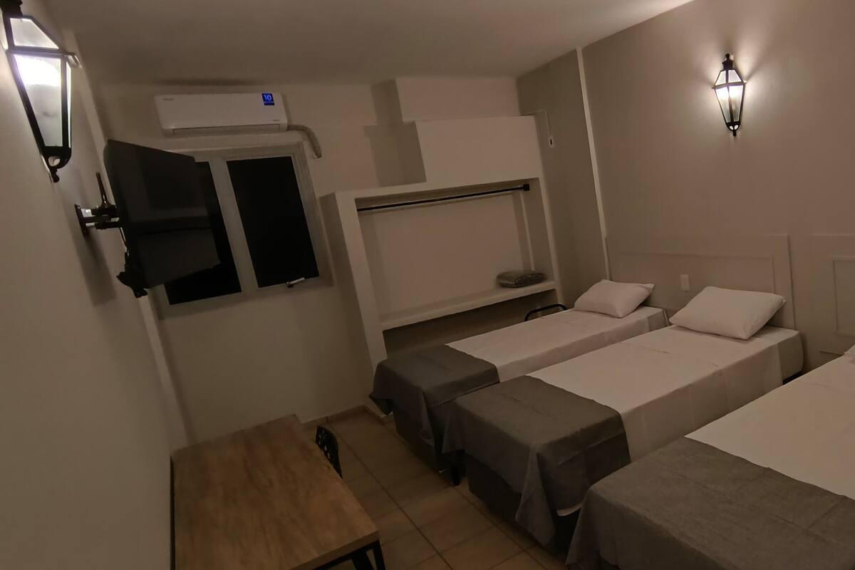 A hotel room with three single beds, a small table, a chair, a wardrobe, and a wall-mounted air conditioner.