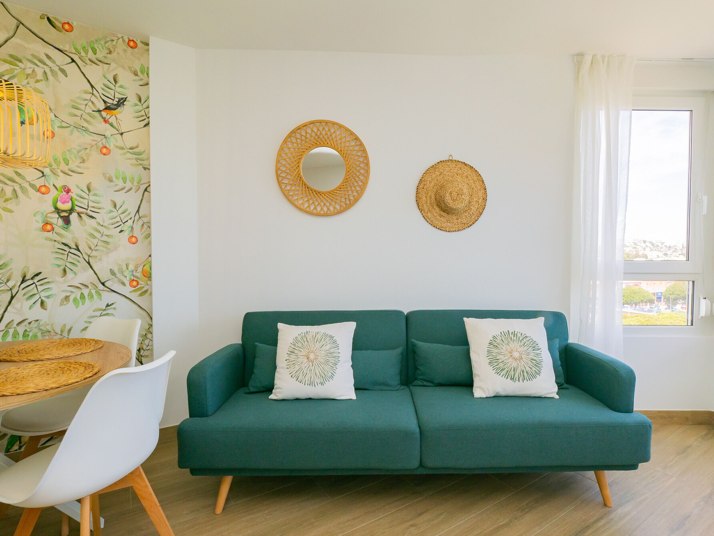 A teal sofa with two decorative pillows, a woven basket on a table, and a wall with a nature-themed mural.