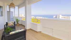 Een balkon met uitzicht op zee, een zithoek met een bank en tafel, en een hanglamp.