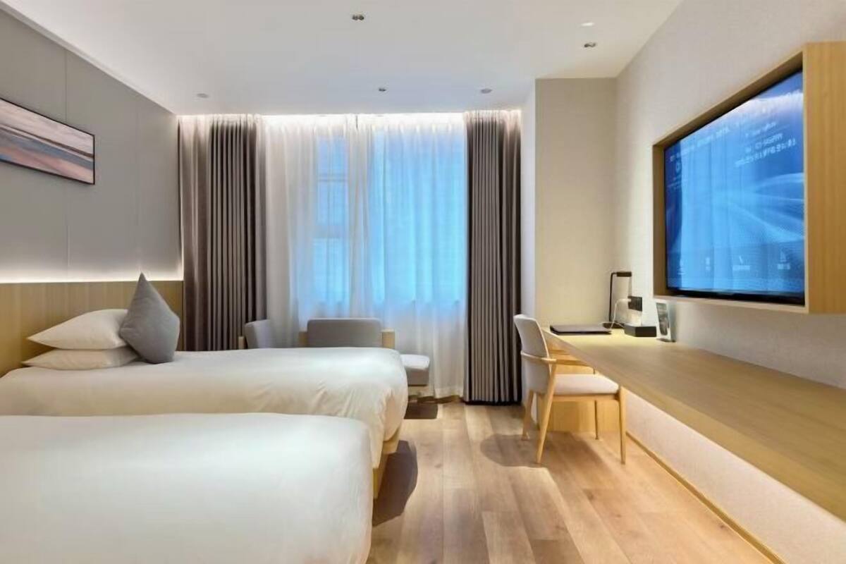 A modern hotel room with two beds, a desk, a chair, a TV, and a large window with curtains.