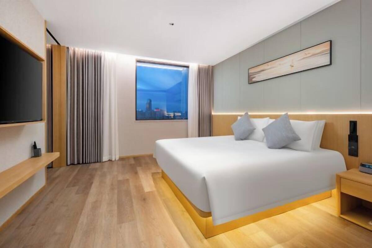 A modern hotel room with a large bed, a TV, a window with a city view, and a wooden floor.