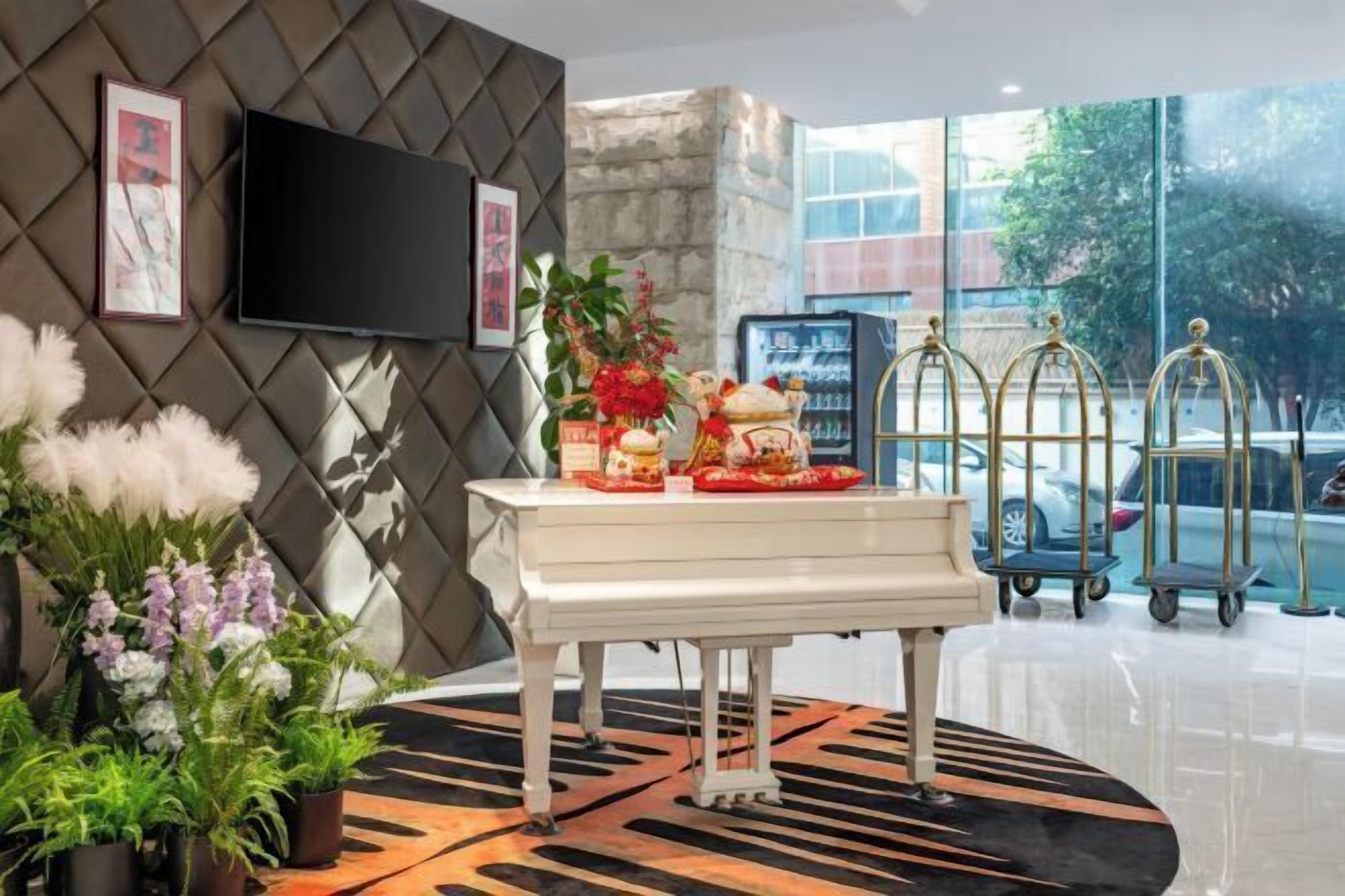 A modern hotel lobby with a white piano, potted plants, a flat-screen TV, and a large window offering a view of the street.
