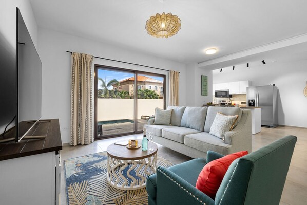 A modern living room with a sofa, a coffee table, and a TV. There is a balcony with curtains and a view of buildings. - Brand NEW 2br2ba w Private Pool in Gated Community (Noord)