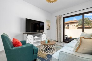A modern living room with a flat-screen TV, teal and light blue sofas, a round coffee table, and a view of a courtyard with palm trees.
