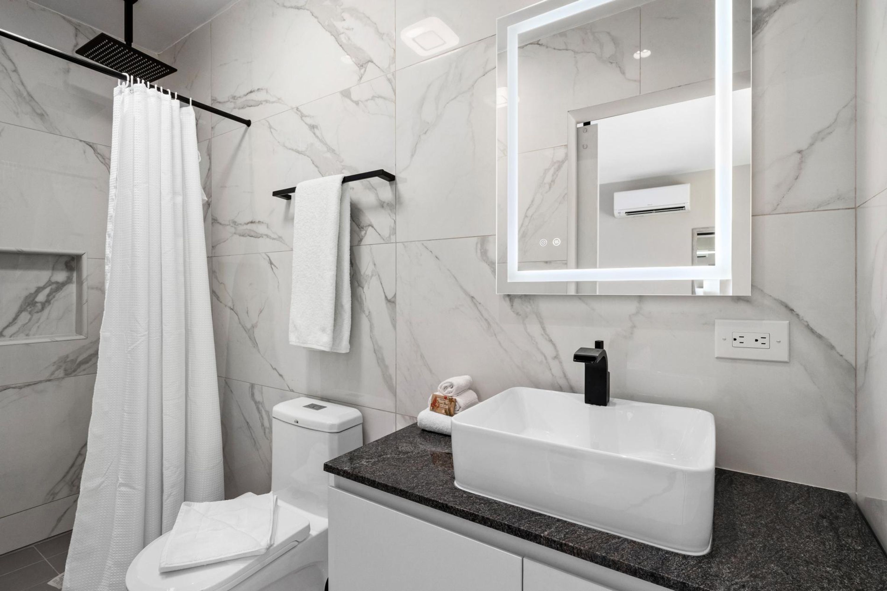 A modern bathroom with a white toilet, a white sink, a shower with a white curtain, and a mirror with a light strip.