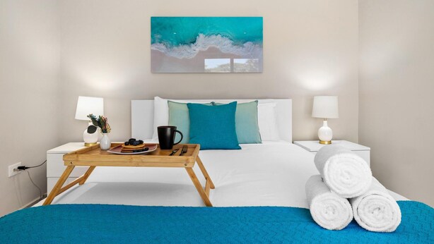 Habitación de hotel con una cama, dos lámparas de noche, un cuadro, una bandeja de madera con alimentos para el desayuno y toallas enrolladas.