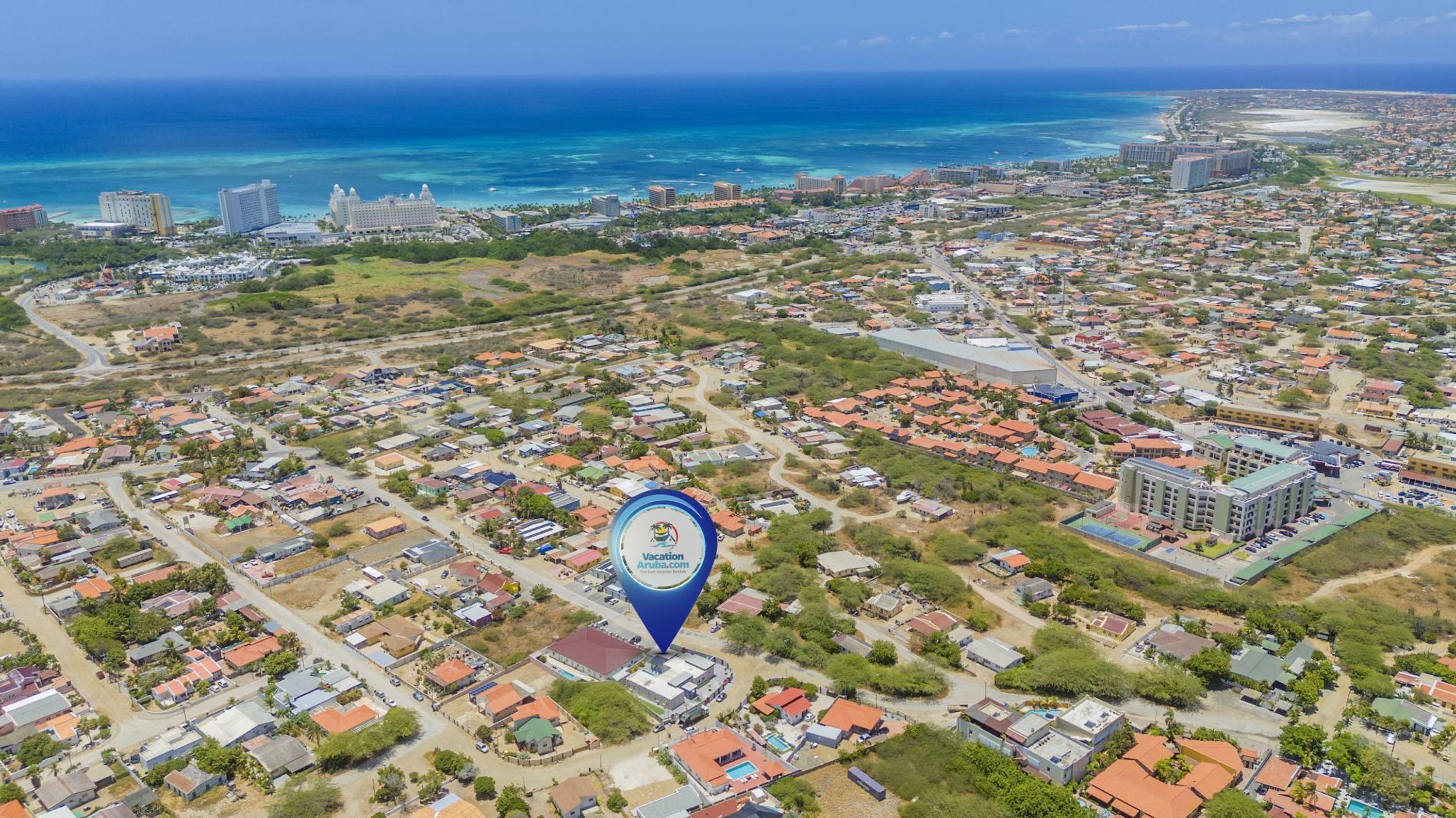 An aerial view of a coastal town with a mix of residential and commercial buildings, a prominent hotel with a large building and a swimming pool, and a clear view of the ocean.