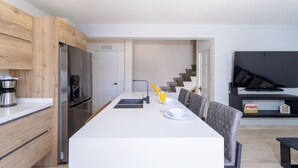 Une cuisine moderne dotée d’un réfrigérateur encastré, d’un coin repas avec une table et des chaises blanches, et d’un téléviseur à écran plat fixé au mur.