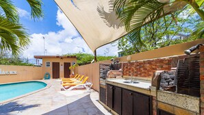 Un espace piscine avec des chaises longues, un barbecue en briques et un auvent ombragé.