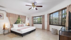 Un dormitorio con cama, ventilador de techo, dos ventanas con cortinas, una mesita de noche con lámpara y un mueble para televisión.