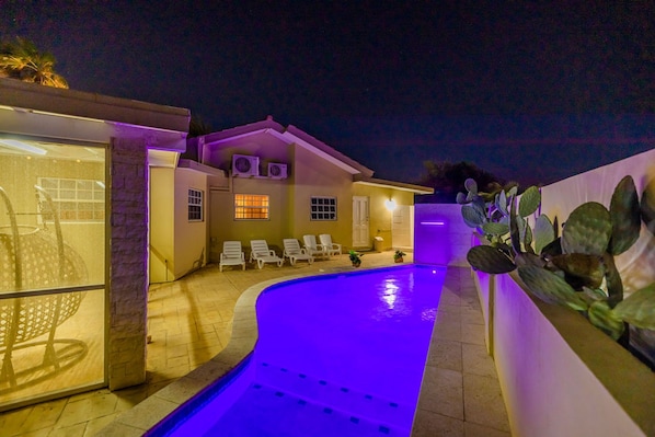 A swimming pool with a curved edge and a blue-lit water feature. There are lounge chairs and a small poolside area with a potted plant.