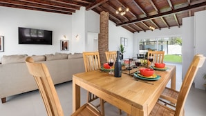A dining area with a wooden table, chairs, and a brick pillar. A television is mounted on the wall, and there is a sofa in the background. - 2BR Countryside Home w Sea Views Private Pool (Noord)