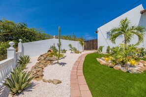 Um jardim bem cuidado com um caminho sinuoso, diversas plantas e um edifício branco com cerca de madeira.