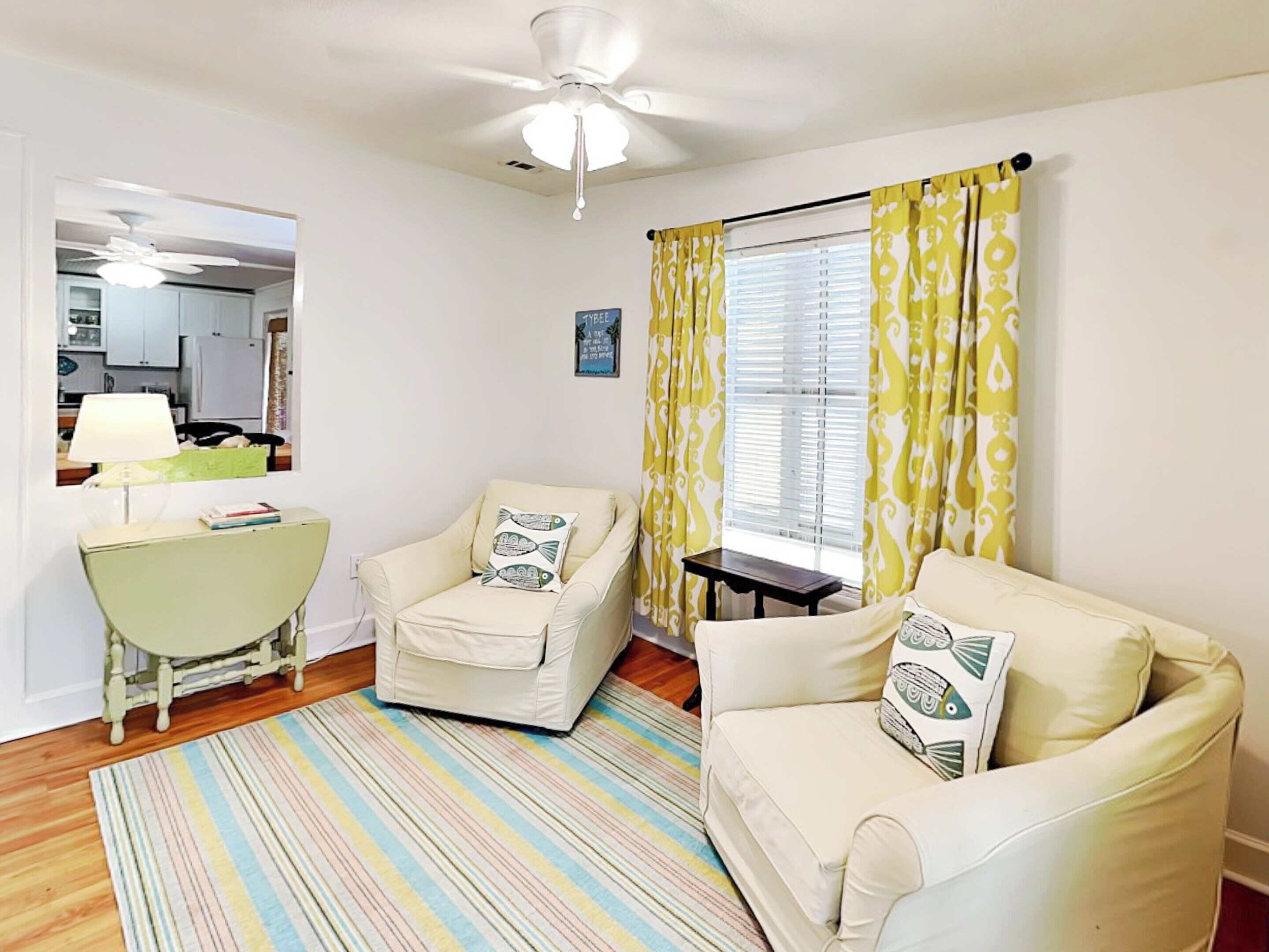 A living room with two cream-colored armchairs, a striped rug, a ceiling fan, and a window with yellow patterned curtains.