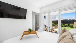 Ein modernes Schlafzimmer mit einem großen Bett, einem Nachttisch aus Holz mit einer Pflanze, einem an der Wand montierten Fernseher und einer Glastür zum Außenbereich mit Terrasse und Blick auf ein Gebäude.