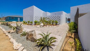 Ein modernes Gebäude mit einer geometrischen Fassade, einem gepflasterten Eingangsbereich und einem angelegten Garten mit Steinen und Pflanzen.