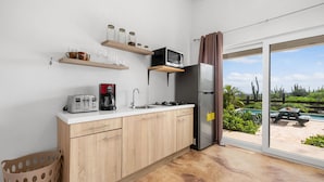 A modern kitchen with wooden cabinets, a microwave, a coffee maker, and a refrigerator. There are shelves above the counter holding jars and glasses. A sliding glass door leads to an outdoor area with a pool and patio furniture. - Breezy Cozy Retreat Arikok National Park Pool (Santa Cruz)