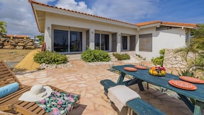 A modern house with a patio area featuring a stone floor, a wooden table with orange plates, and a lounge chair. - Breezy Cozy Retreat Arikok National Park Pool (Santa Cruz)