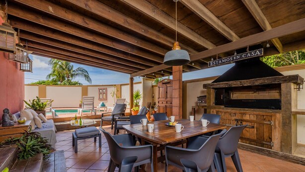 An outdoor patio with a wooden table and chairs, a barbecue grill, and a sign that reads "ESTAMOS EN CASA".