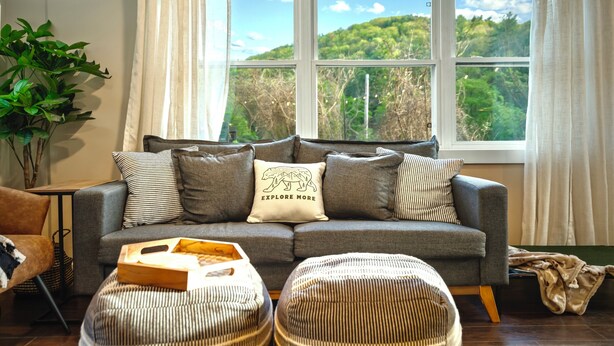 A modern living room with a grey sofa, striped ottoman, and a view of a green landscape through large windows. - Creekside Cabin: Hot Tub, Cinema, EV Charger, Dogs (Luray)