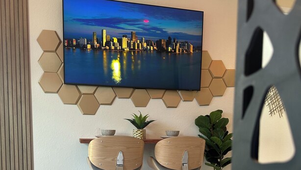 A modern bedroom with a cityscape TV, hexagonal wall decor, two wooden chairs, a bed, and a potted plant. - Tropical Oasis (Quail heights)