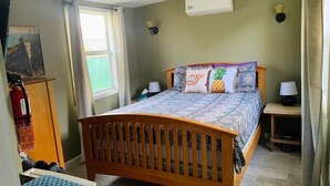 A bedroom with a wooden bed, patterned bedding, two bedside lamps, a nightstand, a window with curtains, a wall-mounted air conditioner, and a painting on the wall. - Country Road, Take me Home- your own Island Cottage! (Savannah)