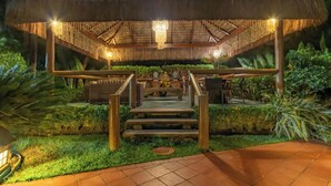 A covered outdoor seating area with a thatched roof, steps leading up to it, and surrounding greenery. - Hotel Coral Beach  (Tamandaré)