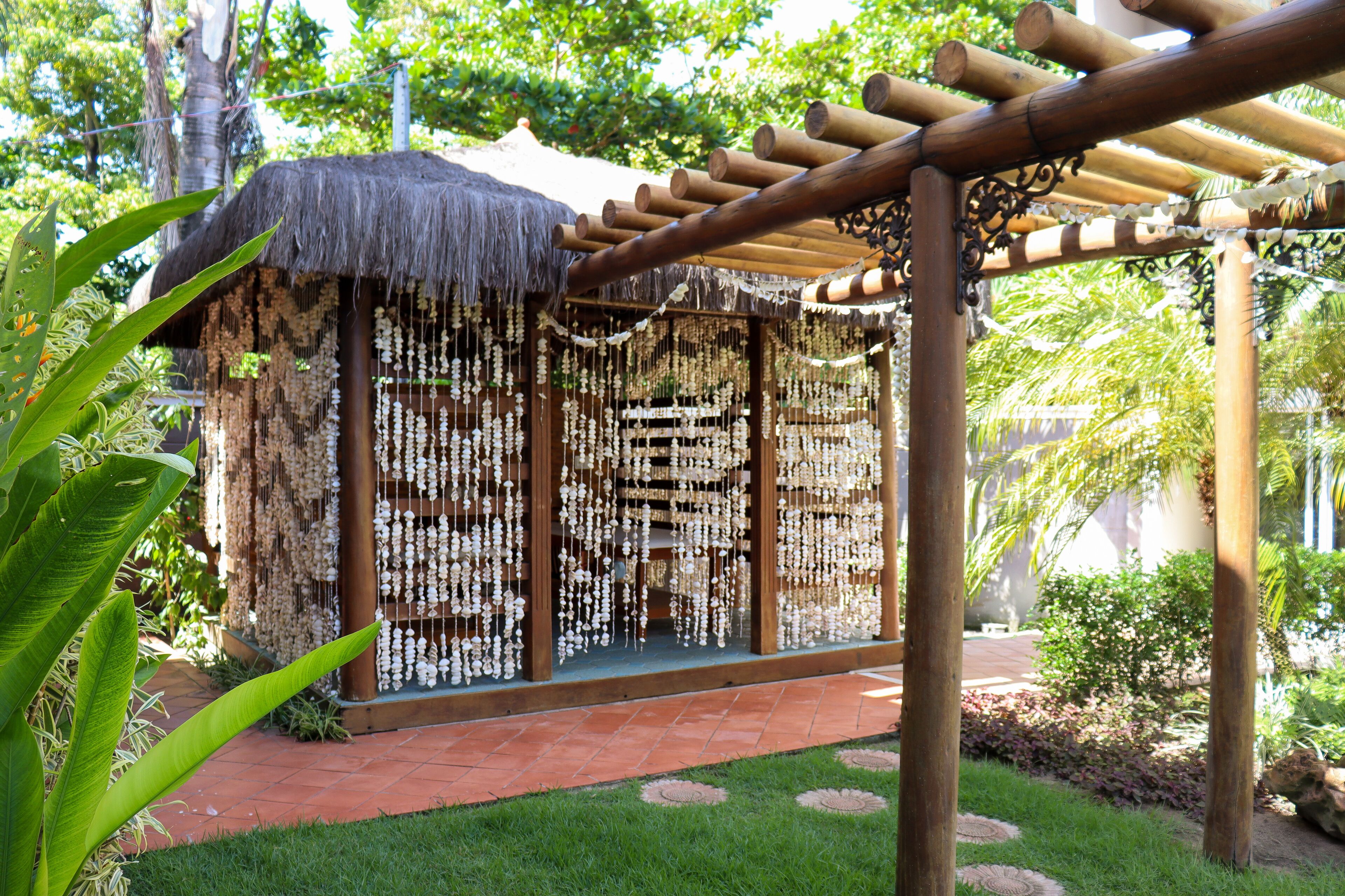Een pergola van bamboe met rieten dak, omgeven door weelderige begroeiing en een verzorgde tuin.