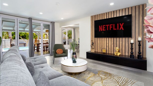 Un salon moderne doté d’un grand téléviseur à écran plat fixé sur un mur en bois, d’un canapé d’angle gris, d’une table basse ronde et d’une porte en verre donnant sur un espace extérieur avec une piscine.