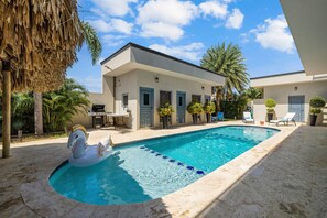 A swimming pool with a decorative swan float, a patio area with lounge chairs, and a building with a modern architectural style. - Magical Oasis 6BR w Large Pool Gazebo Views (Noord)