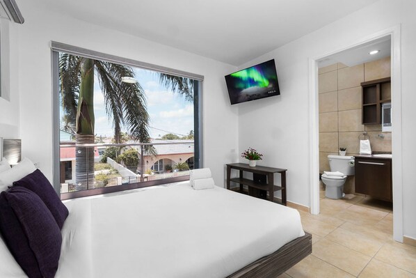 A modern hotel room with a large bed, a TV mounted on the wall, a balcony with a view of palm trees, and a separate bathroom area. - Magical Oasis 6BR w Large Pool Gazebo Views (Noord)