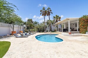 A backyard with a swimming pool, lounge chairs, palm trees, and a covered patio area.