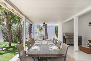 A covered outdoor dining area with a long table set for six, surrounded by chairs, a grill, and a view of a garden with palm trees. - 4br4ba w Private Pool Walking Distance to Ritz (Noord)