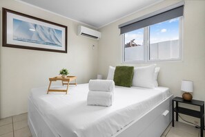 A neatly arranged bedroom with a bed, a small table with a plant, a window with a view, and an air conditioning unit.